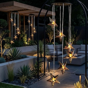 Może przedstawiać: Zestaw wiszących lamp solarnych do użytku zewnętrznego, składający się z wielu metalowych lampionów w kształcie gwiazd z ażurowymi wzorami. Każda gwiazda świeci ciepłym światłem i jest zawieszona na białych sznurkach.