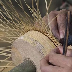 May include: Close-up of a craftsperson weaving a basket. The basket is made of light brown, thin strips of wood. The craftsperson is using a wooden tool to weave the strips together. The basket is in the early stages of construction.