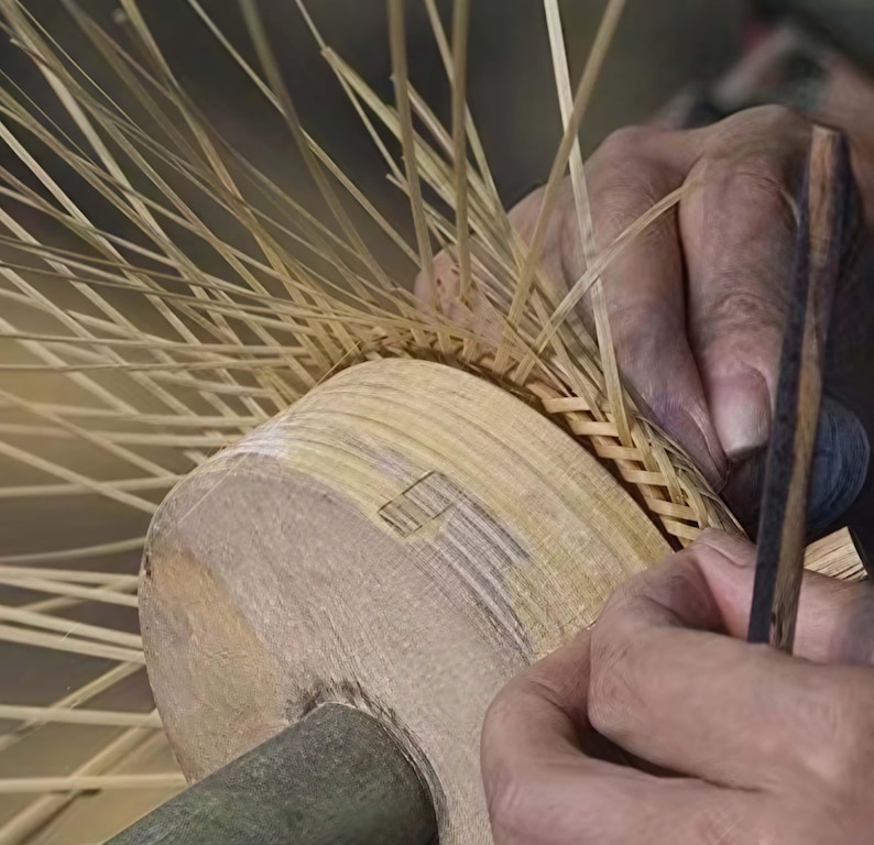 May include: Close-up of a craftsperson weaving a basket. The basket is made of light brown, thin strips of wood. The craftsperson is using a wooden tool to weave the strips together. The basket is in the early stages of construction.