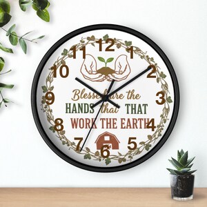 Könnte beinhalten: Eine runde Wanduhr mit schwarzem Rahmen. Das Zifferblatt zeigt den Text "Blessed are the hands that work the earth" und eine Grafik von Händen, die einen Spross halten. Die Zahlen sind braun.