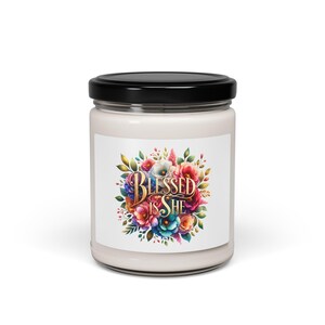 Pode incluir: Uma vela branca num frasco de vidro com tampa preta. O rótulo apresenta um desenho floral com as palavras "BLESSED IS SHE" em letras douradas. As flores são em tons de vermelho, azul, rosa e amarelo.