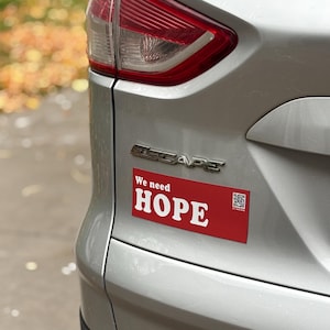 May include: A silver SUV with a red bumper sticker that reads "We need HOPE" in white capital letters. The car's taillight is visible, and the word "ESCAPE" is displayed on the vehicle.