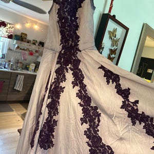 May include: A sleeveless, floor-length gown in a light purple color with intricate black lace detailing. The lace is arranged in a geometric pattern, creating a striking contrast against the soft fabric.