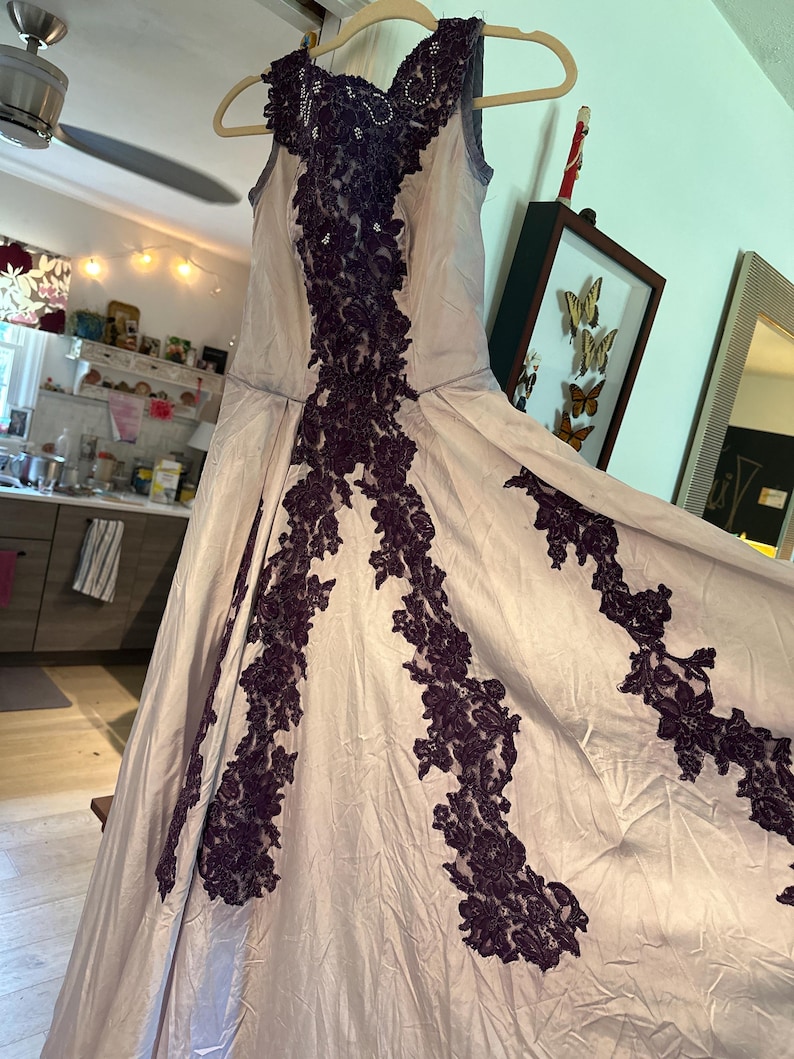 May include: A sleeveless, floor-length gown in a light purple color with intricate black lace detailing. The lace is arranged in a geometric pattern, creating a striking contrast against the soft fabric.