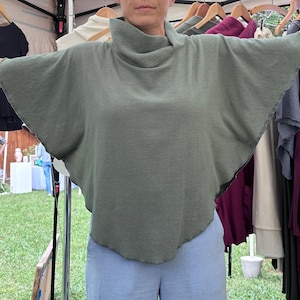May include: A sage green poncho-style top with a high, rolled neckline and raw, unfinished edges. The top has a wide, flowing shape with short sleeves. Other clothing items are visible in the background.