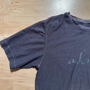 May include: A dark gray t-shirt with a white stitched seam around the neckline and sleeves. The shirt has the word "alive" printed on the front in a white, cursive font.