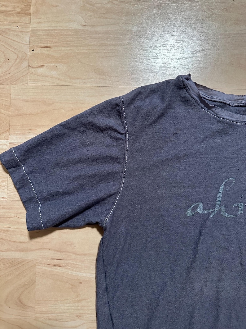 May include: A dark gray t-shirt with a white stitched seam around the neckline and sleeves. The shirt has the word "alive" printed on the front in a white, cursive font.