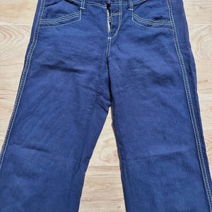 May include: Dark blue denim pants with white stitching detail. The pants have a button and zipper closure.