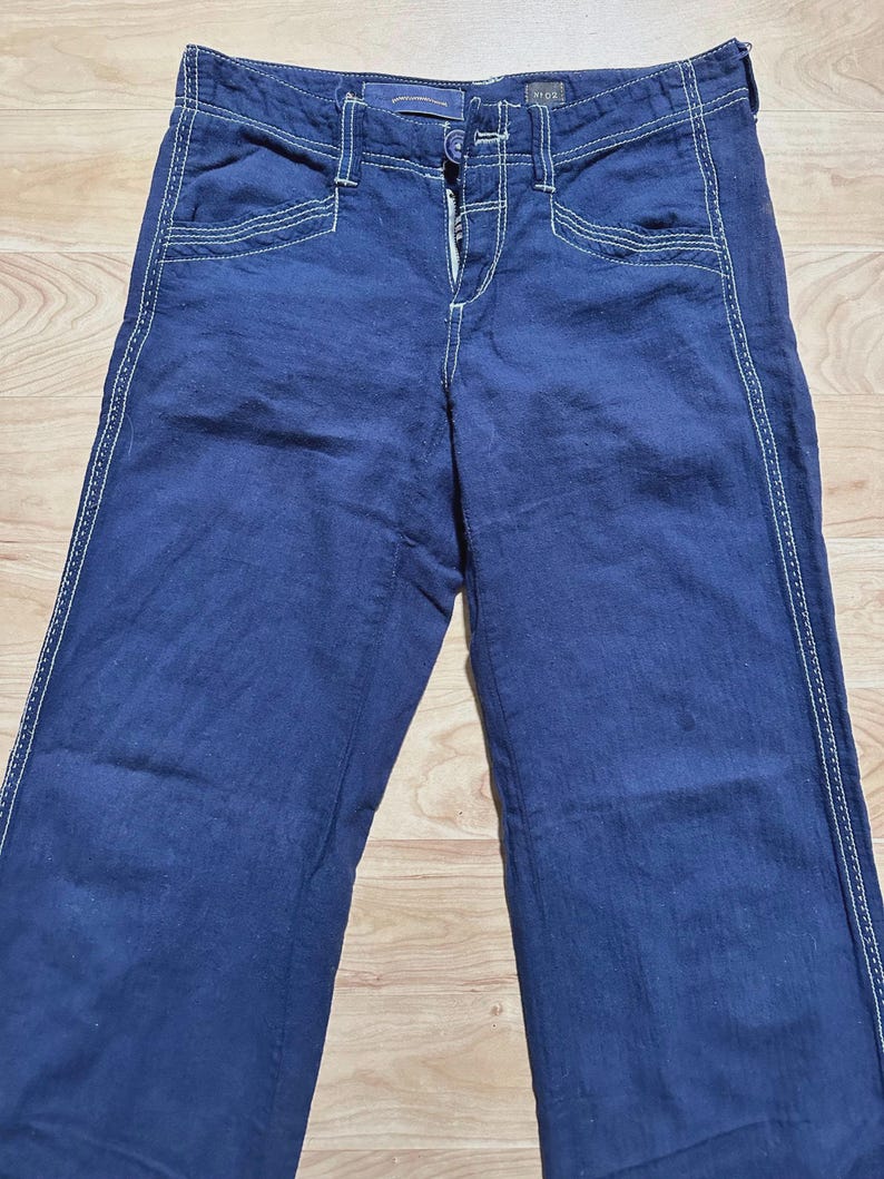 May include: Dark blue denim pants with white stitching detail. The pants have a button and zipper closure.