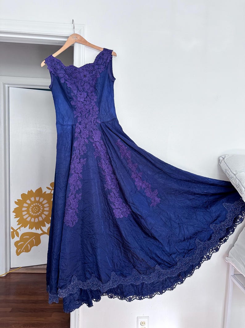 May include: A long, blue, sleeveless dress with purple lace detailing. The dress has a scoop neckline and a full skirt.