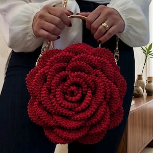 May include: A red crocheted handbag shaped like a rose. The bag has a gold chain strap and a gold handle. The bag is held by a person wearing a white blouse and dark pants. The bag is a unique accessory.
