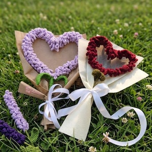 Könnte beinhalten: Zwei Sträuße gehäkelter Blumen auf Gras. Einer hat ein lavendelfarbenes Herz, der andere ein burgunderfarbenes Herz. Beide sind in Papier verpackt und mit weißen Bändern gebunden. Zusätzliche gehäkelte Lavendel- und grüne Stiele sind ebenfalls sichtbar.