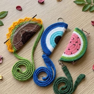 May include: Three crochet eyeglass cases: a taco, an evil eye, and a watermelon slice. The taco case is brown with yellow and green trim. The evil eye case is blue, white, and black. The watermelon case is pink, green, and white.