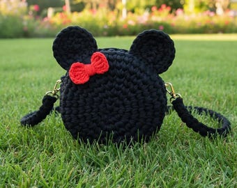 May include: A black crochet handbag shaped like a mouse, with two large ears and a red bow. The bag has a round shape and a black strap. The bag is sitting on green grass.