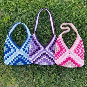 Può includere: Tre borse all'uncinetto fatte a mano nei toni del blu, viola e rosa. Ogni borsa presenta un design a granny square e un manico a contrasto. Le borse sono esposte su erba verde, mostrando i loro colori e le loro trame vivaci.
