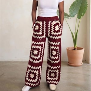 May include: Wide-leg trousers crafted from crochet yarn in a deep burgundy and cream color scheme. The pants feature a repeating granny square pattern throughout, a high-waisted fit with an elasticized waistband, and two side pockets.