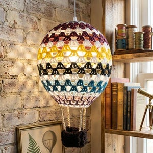 Könnte beinhalten: Gehäkelte Heißluftballon-Hängelampe mit einem schwarzen Korb. Die Lampe ist an einem weißen Kabel aufgehängt und hat farbige Streifen in Weiß, Gelb, Schwarz, Blau und Burgunderrot. Der Hintergrund ist eine Backsteinmauer.