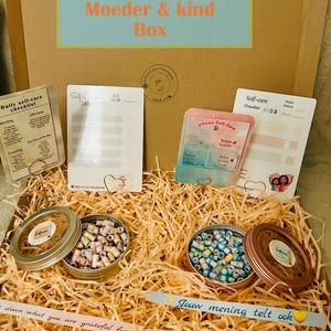 Op de afbeelding: Een geschenkdoos met de tekst "Moeder & kind Box" bevat zelfzorgchecklists, kleine blikken gevuld met kleurrijke papieren kralen en decoratieve kaarten. De doos is gevuld met stro en heeft handgeschreven notities.