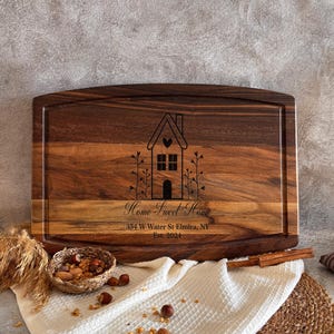 May include: A rectangular dark brown wooden cutting board with a grooved border. The center features a laser-engraved graphic of a house with a heart, the text "Home Sweet Home", the address "354 W Water St Elmira, NY", and "Est. 2024".