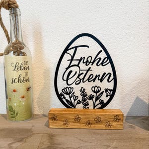 Könnte beinhalten: Schwarze Osterdekoration in Eiform mit der Aufschrift "Frohe Ostern" und floralen Details, auf einem Holzsockel. Eine Glasflasche mit dem Text "Das Leben ist schön" ist ebenfalls zu sehen.