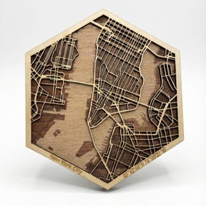 Può includere: Mappa in legno esagonale di New York City, con dettagliati layout di strade e corsi d'acqua. La mappa è tagliata al laser con una cornice in legno chiaro e un interno in legno più scuro, con il testo "New York City" e coordinate.