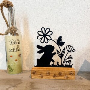 Könnte beinhalten: Schwarze Metall-Silhouette eines Hasen mit Blumen- und Schmetterlingsdesign, auf einem Holzsockel mit Blumenschnitzereien. Eine Glasflasche mit dem Text "Das Leben ist schön" und einem Seilgriff ist ebenfalls im Bild.