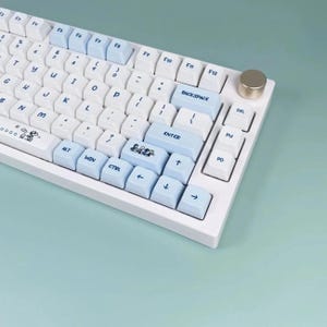 May include: White mechanical keyboard with light blue and white keycaps. The keyboard features a gold-colored knob and a cartoon graphic on the space bar. The keys are arranged in a standard QWERTY layout.