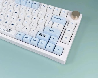 May include: White mechanical keyboard with light blue and white keycaps. The keyboard features a gold-colored knob and a cartoon graphic on the space bar. The keys are arranged in a standard QWERTY layout.