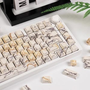 May include: A white mechanical keyboard with keycaps featuring a marble design in shades of cream, gray, and brown. The keyboard has a white frame and a round white knob. Several loose keycaps are scattered around the keyboard.