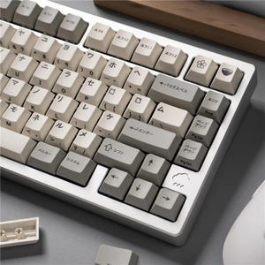 May include: A white and gray mechanical keyboard with Japanese characters and symbols on the keycaps. The keyboard has a minimalist design with a white frame and gray accent keys. The keycaps have a retro aesthetic.