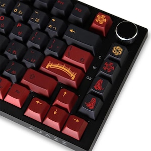 May include: Black mechanical keyboard with red and black keycaps. Some keycaps feature Japanese characters and decorative symbols. The keyboard has a volume control knob and the brand name "AJAZZ" is visible.