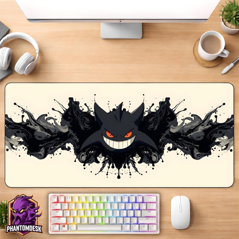 May include: A rectangular desk mat with a black cartoon character with a wide grin and red eyes, set against a backdrop of black ink-like splatters. A white keyboard and mouse are visible below the mat. The PhantomDesk logo is in the lower left corner.
