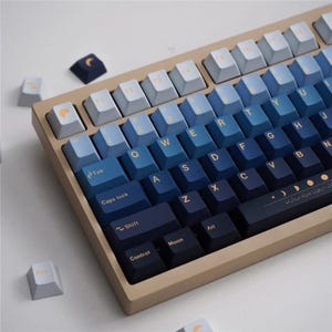 May include: A beige mechanical keyboard with keycaps in a gradient of blue hues. The keycaps feature white lettering and symbols, with some keys displaying moon and sun icons. Additional loose keycaps are scattered around the keyboard.