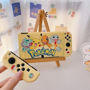 Könnte beinhalten: Gelbes Nintendo Switch-Gehäuse und Controller mit Pokémon-Motiv. Das Gehäuse zeigt Bulbasaur, Pikachu, Charmander und Squirtle. Das Wort "Pokémon" ist in Blau und Gelb geschrieben. Der Controller hat schwarze Knöpfe und ein rot-weißes Pokéball-Design.