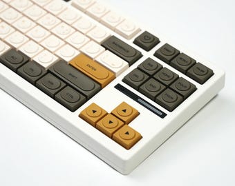 May include: A white mechanical keyboard with a variety of keycap colors, including cream, olive green, and mustard yellow. The keyboard has a compact layout with function keys and arrow keys. The keycaps are a mix of colors and shapes.
