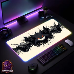 May include: A cream-coloured gaming mouse mat featuring a black cartoon character with a wide grin and red eyes, surrounded by black ink-like splatters. The mat has a colourful LED edge and the PhantomDesk logo.