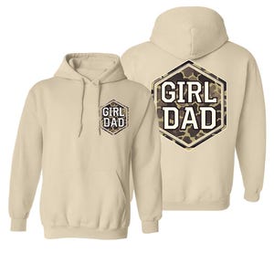 May include: Two beige hoodies with the text "GIRL DAD" in a camouflage design. One hoodie has the text on the front, and the other has the text on the back. Both hoodies have a hood and a front pocket.