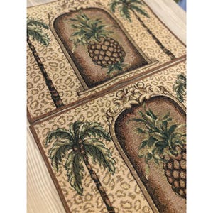 Puede incluir: Dos manteles individuales rectangulares con temática tropical. Cada uno presenta un diseño de tapiz detallado de una piña dentro de un marco arqueado, flanqueado por palmeras. El fondo tiene un estampado de leopardo beige. Los manteles individuales están ribeteados en marrón.