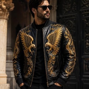 May include: A black bomber jacket featuring intricate gold embroidery of eagles on the chest and ornate scrollwork patterns extending down the sleeves. The jacket has a ribbed collar, a front zipper closure, and is worn over a black shirt.