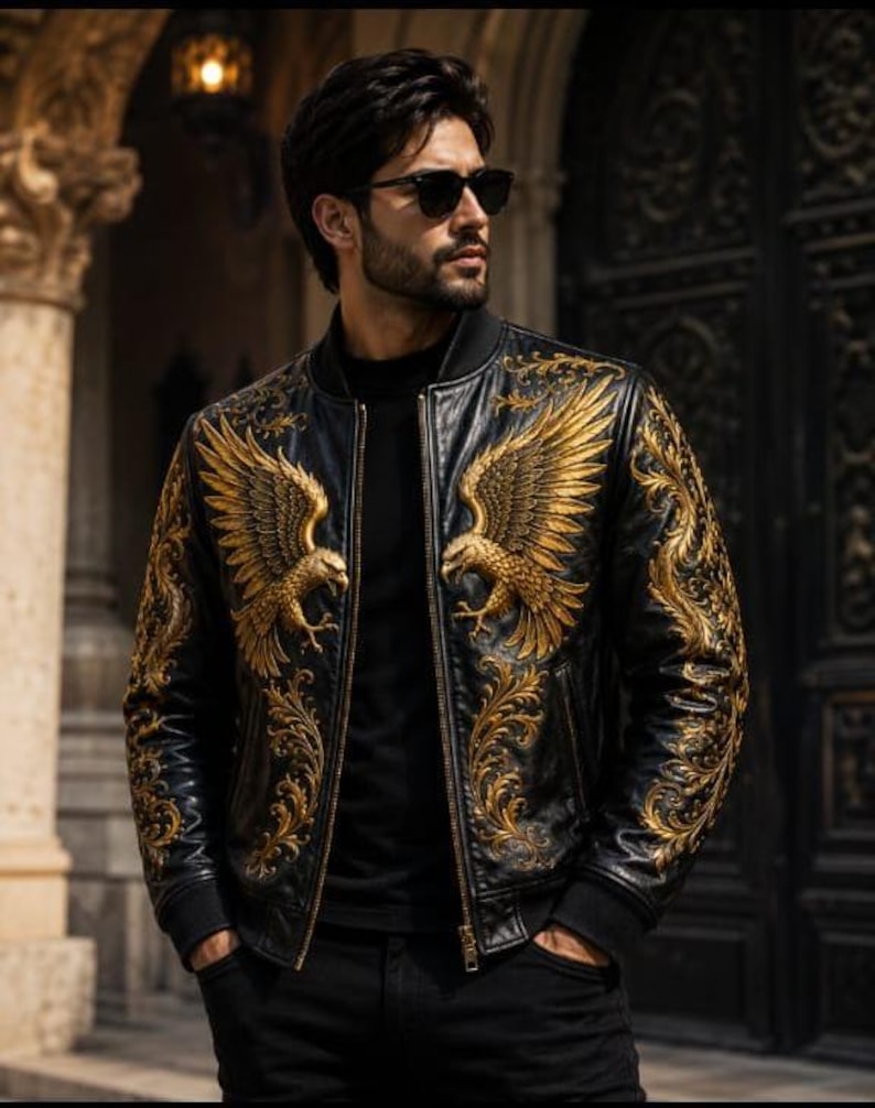 May include: A black bomber jacket featuring intricate gold embroidery of eagles on the chest and ornate scrollwork patterns extending down the sleeves. The jacket has a ribbed collar, a front zipper closure, and is worn over a black shirt.