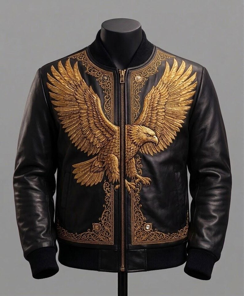May include: A black leather bomber jacket featuring a large, detailed gold-colored eagle emblem across the chest. The design includes intricate gold filigree patterns along the front panels and a central gold-toned zipper closure with ribbed cuffs.