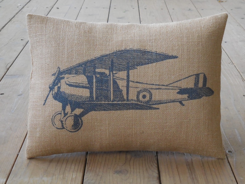 May include: A burlap pillow with a vintage airplane design printed in black. The airplane is a biplane with a propeller and a single engine.