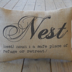 May include: A burlap pillow with the word "Nest" in a decorative script font. The pillow also has the definition of "nest" printed below the word: "(nest) noun: a safe place of refuge or retreat."