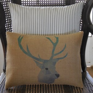 May include: A brown burlap cushion with a blue and green deer silhouette design. The cushion is sitting on a black wicker chair with a white and grey striped cushion behind it.