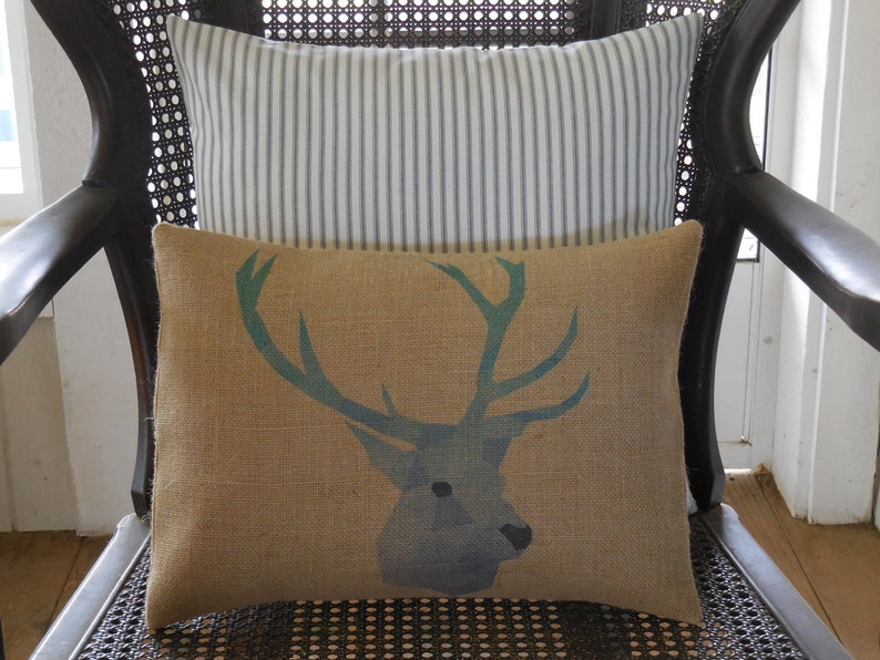 May include: A brown burlap cushion with a blue and green deer silhouette design. The cushion is sitting on a black wicker chair with a white and grey striped cushion behind it.