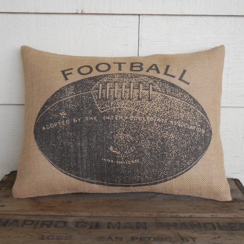 May include: A brown burlap pillow with a black and white image of a football. The text "FOOTBALL" is printed above the football and the text "ADOPTED BY THE INTERCOLLEGIATE FOOTBALL ASSOCIATION" is printed below the football.