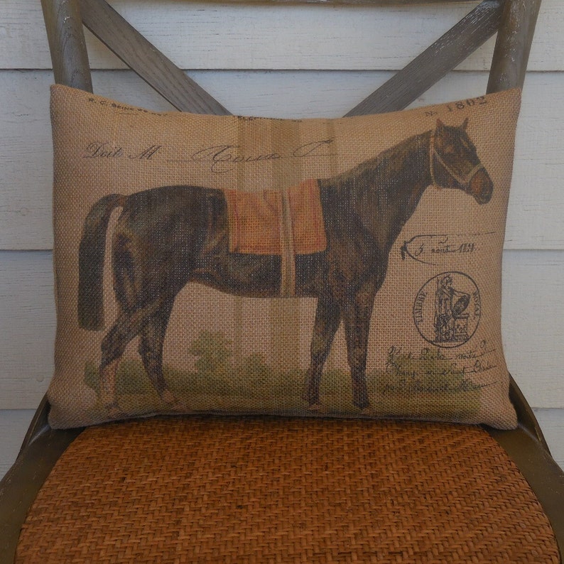 Op de afbeelding: Een bruin jute kussen met een vintage illustratie van een paard met een zadel. Het kussen heeft een bruin-wit gestreepte rand en de tekst "Date M. Gouve" staat bovenaan gedrukt.