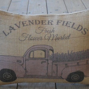 May include: A burlap pillow with a vintage truck design. The truck is light purple and has a bed full of lavender. The pillow has the text "Lavender Fields Fresh Flower Market" printed on it.