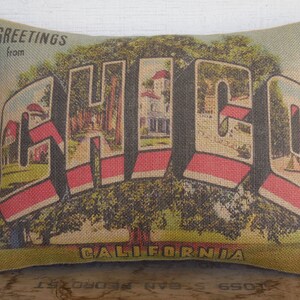 May include: A burlap pillow with a vintage postcard design featuring the text "Greetings from CALI FORNIA" and a stylized image of a city with trees and buildings.