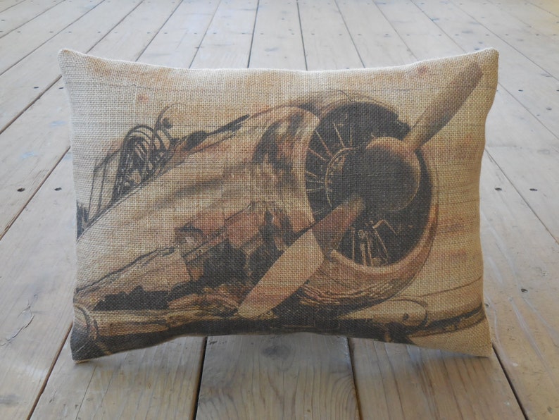 May include: A brown and beige burlap pillow with a vintage airplane printed on it. The airplane has a propeller and is in flight.